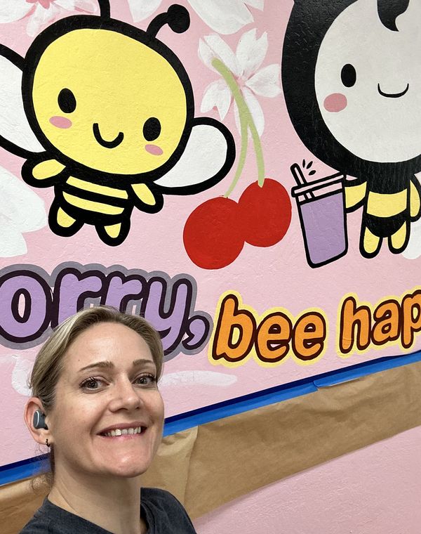 Kelli in front of a pink cafe mural she painted with cartoon bees, cherries and a boba drink