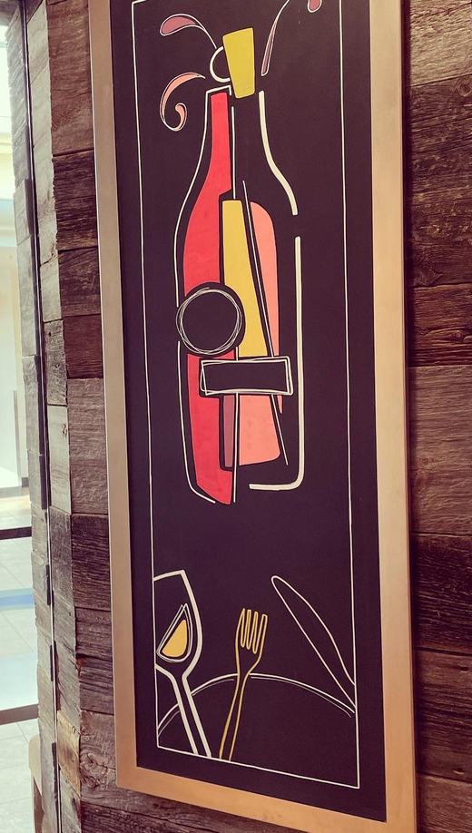 Painted wine bottle and glass artwork in a gold frame on a wood column